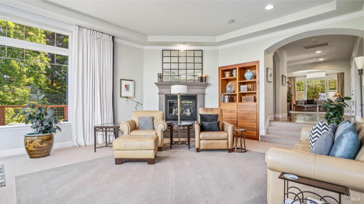 Living room featuring a fireplace with a mirror above, beige armchairs, and a large window offering a view of greenery.