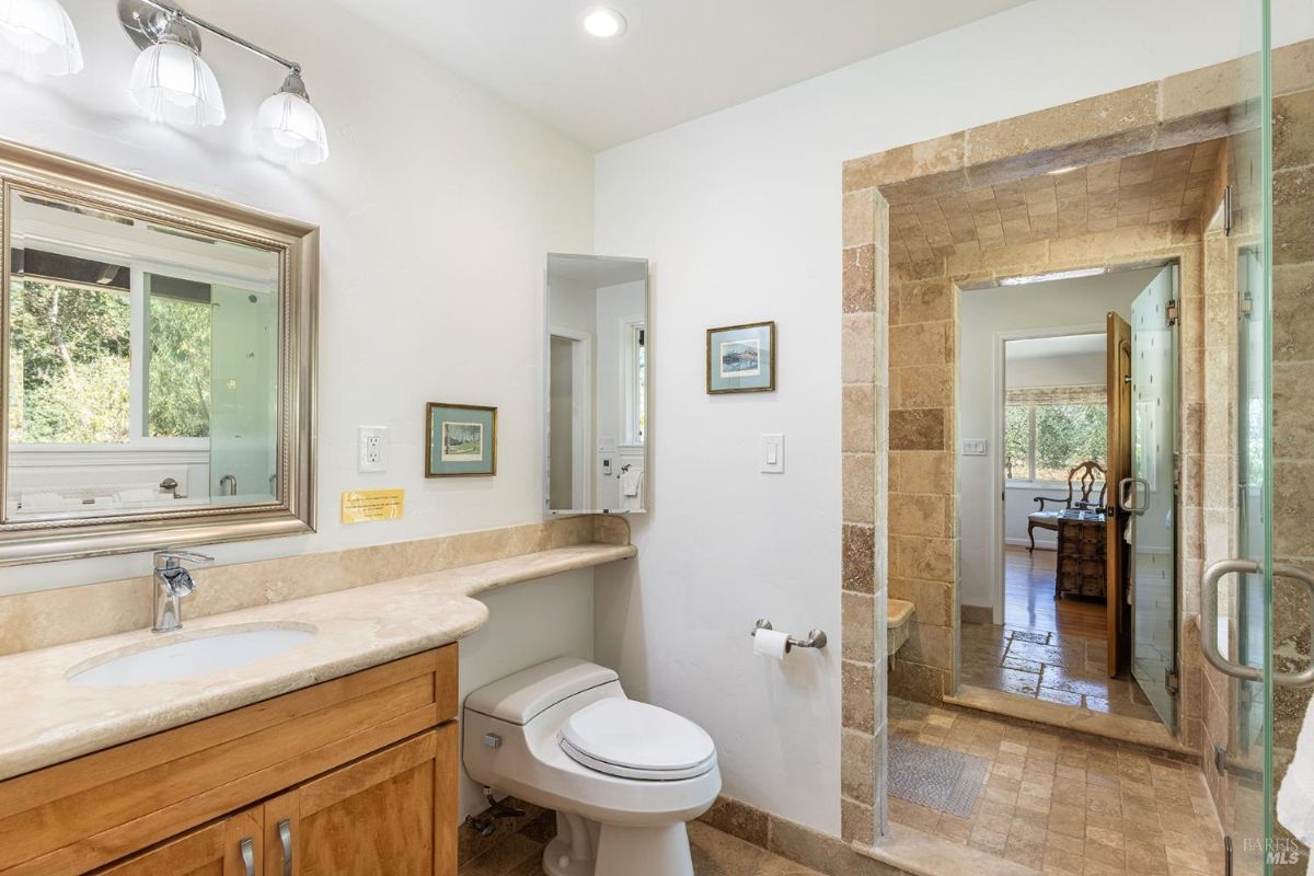 Bathroom with beige countertops, a toilet, a glass-enclosed shower, and a view of an adjacent bedroom.