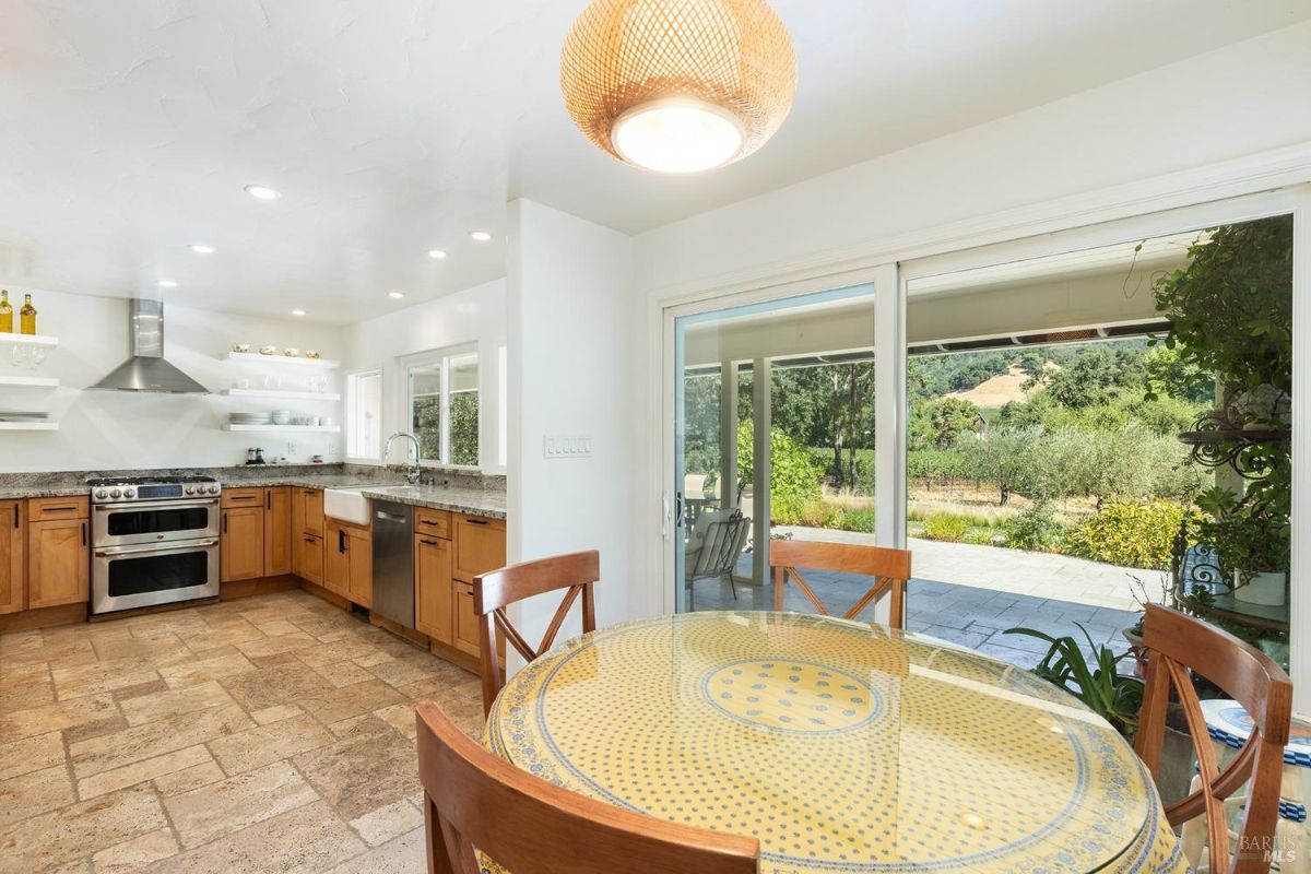 Bright kitchen with wooden cabinets, granite countertops, open shelving, and a dining area with a yellow-patterned table.