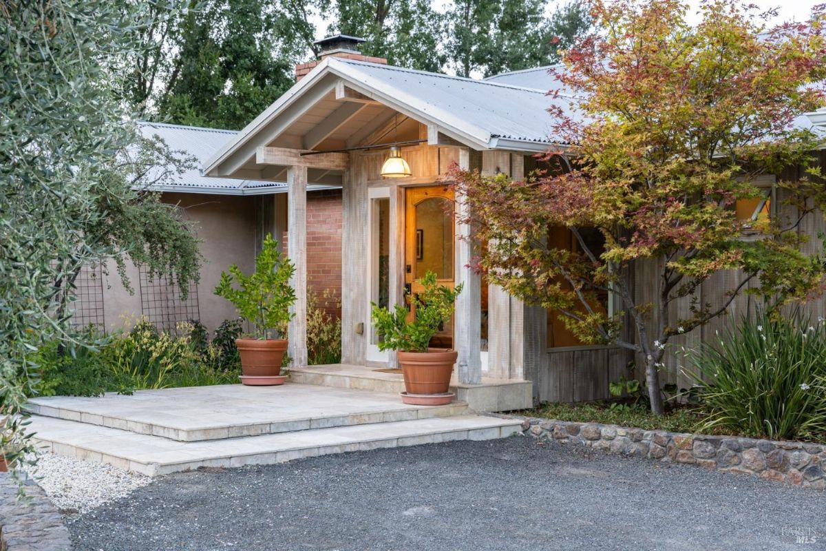 Front entrance displays a covered porch with potted plants and a pathway surrounded by trees.