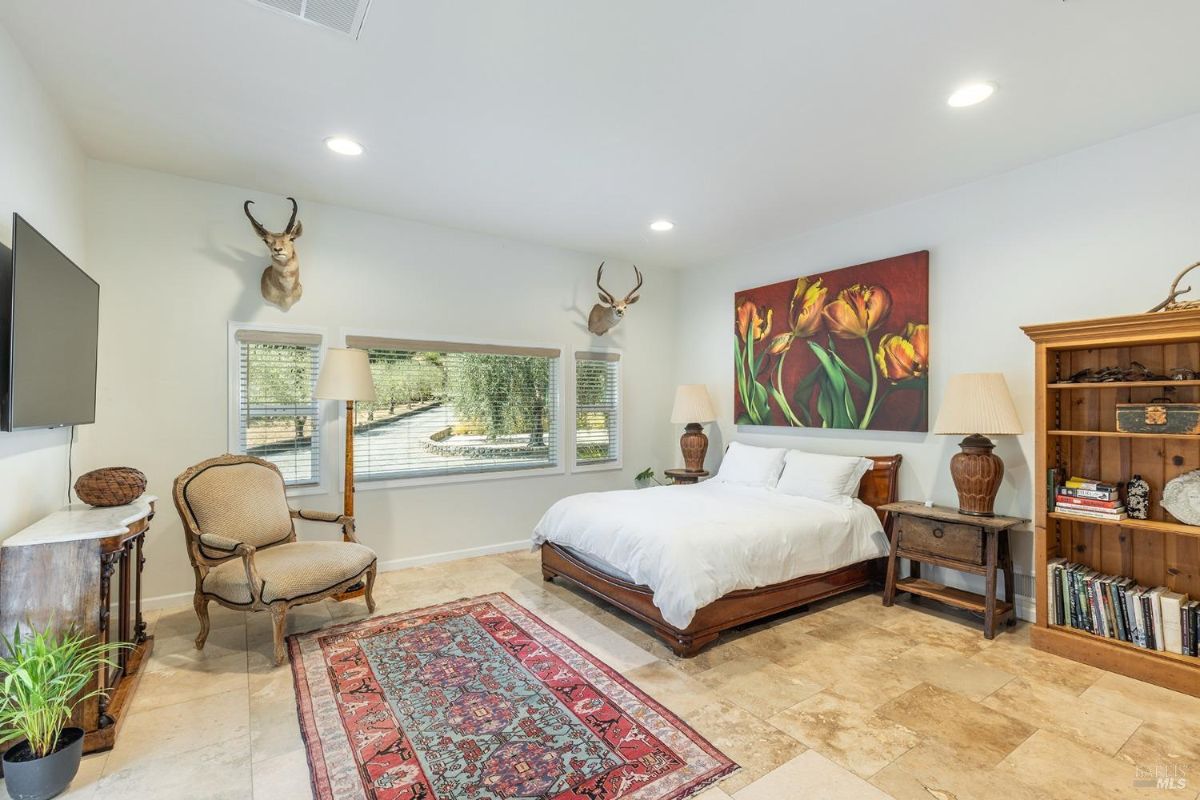 Bedroom with stone tile flooring, a colorful rug, mounted antlers, and a large painting above the bed.