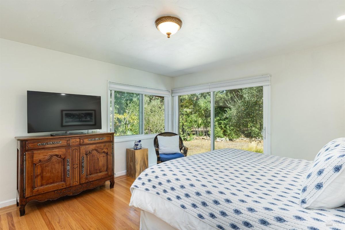 Bedroom with a TV on a wooden cabinet, large windows, and a blue accent chair.