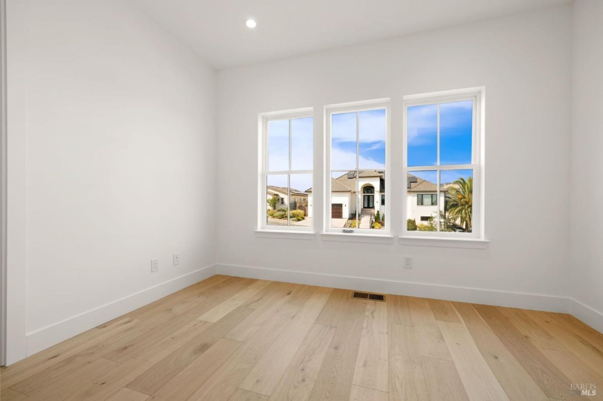 Empty room with hardwood floors and three large windows overlooking neighboring houses.