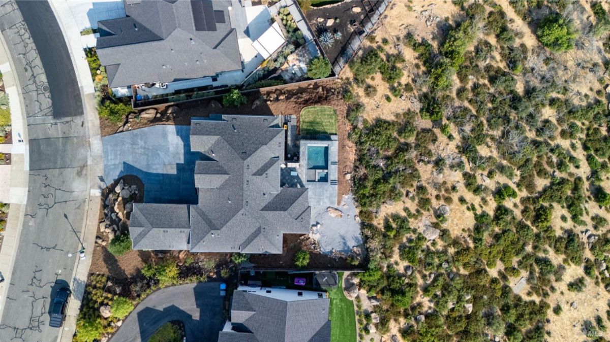 Aerial view of a residential property showing a house, driveway, backyard with a pool, and adjacent open land with vegetation.